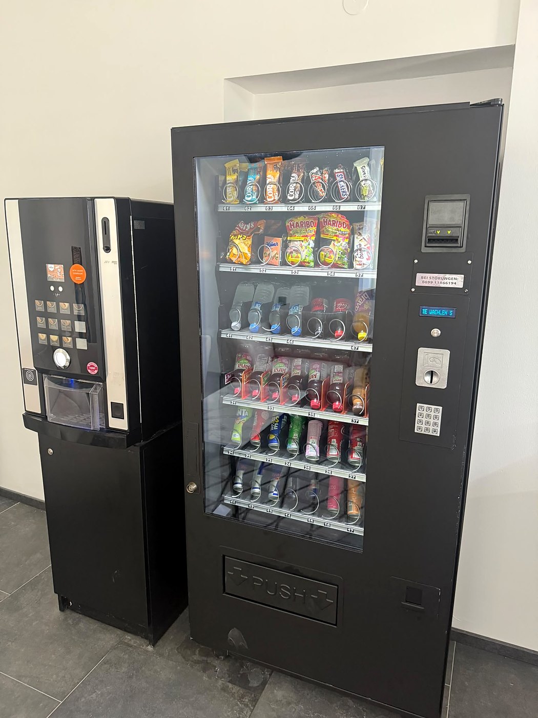 Großer Snack- und Getränkeautomat in einem Gang, daneben ein Kaffeeautomat; Regale mit Riegeln, Chips, Gummibärchen und Flaschen hinter Glas, davor ein Fliesenboden