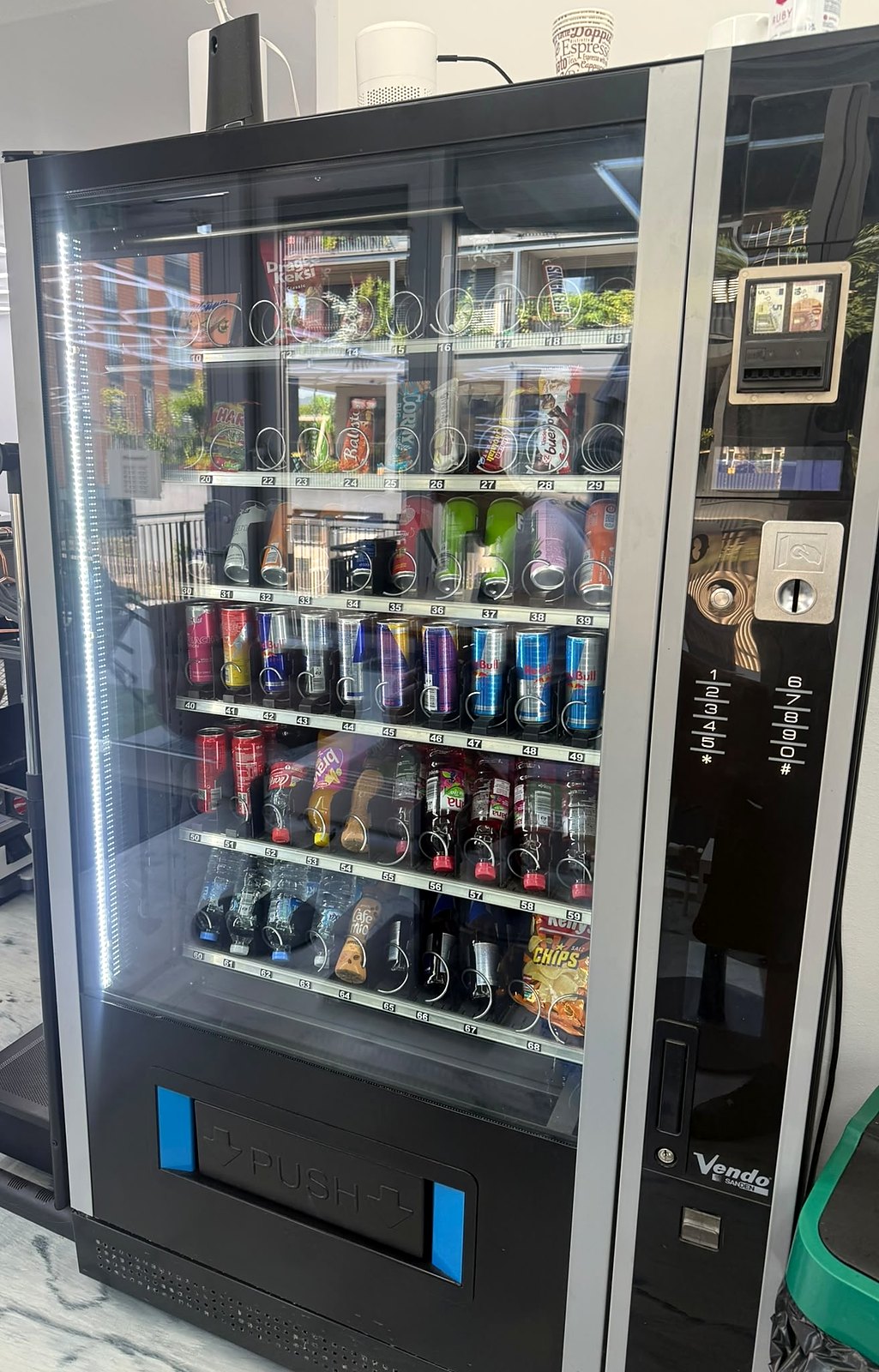 Getränke- und Snackautomat in voller Höhe, aufgestellt in einem Fitnessstudio: beleuchtete Glasfront mit vollen Regalen, rechts die Bedienspalte mit Münzeinwurf und Tastenfeld, darunter Marmorboden